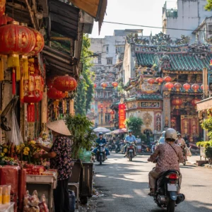 A lively street scene in Saigon’s Cholon Chinatown, with red lanterns hanging outside traditional shops and a motorbike passing through the bustling street market.